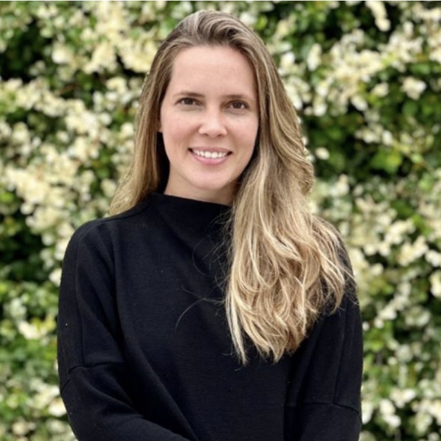 Carol Frigério sorrindo, de blusa preta, em frente a um muro de flores